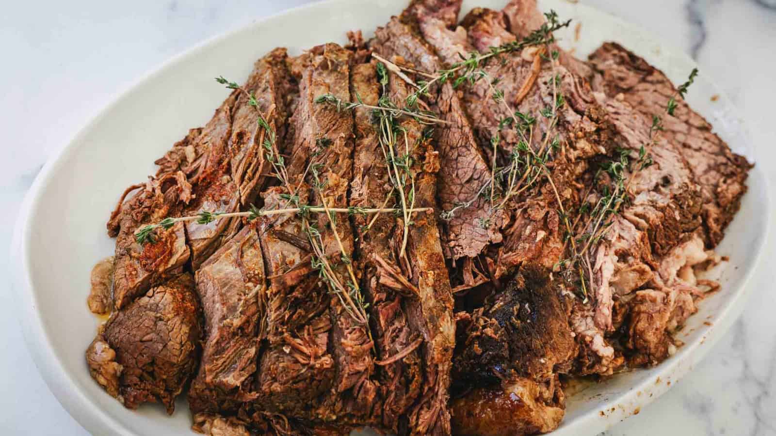 A white oval plate holds several slices of cooked, tender beef roast topped with sprigs of fresh thyme on a light marble surface.