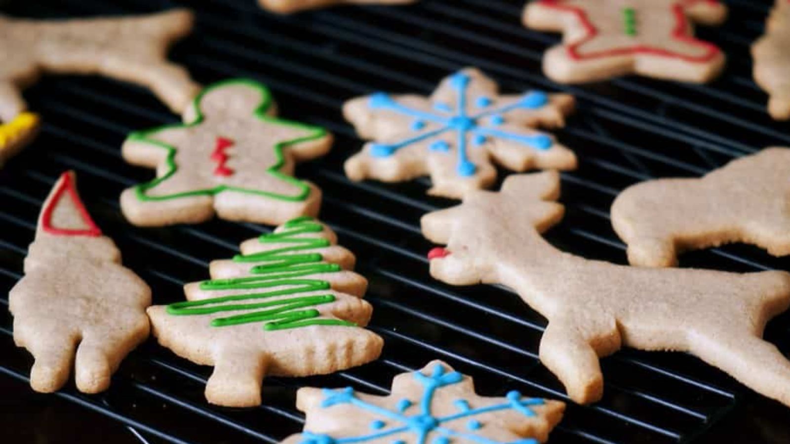 Assorted Christmas-themed sugar cookies, some decorated with colorful icing, cooling on a black wire rack.