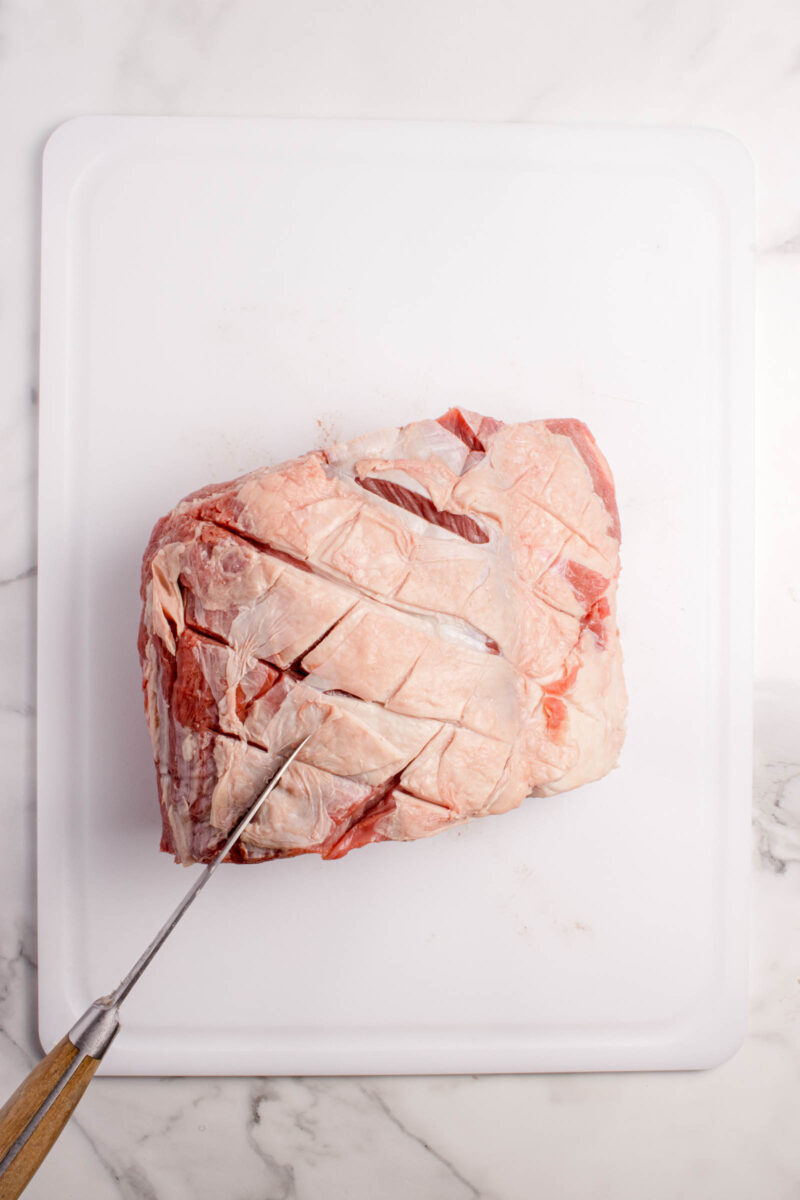 A raw piece of meat with a scored surface is placed on a white cutting board; a knife is making diagonal cuts on the meat.