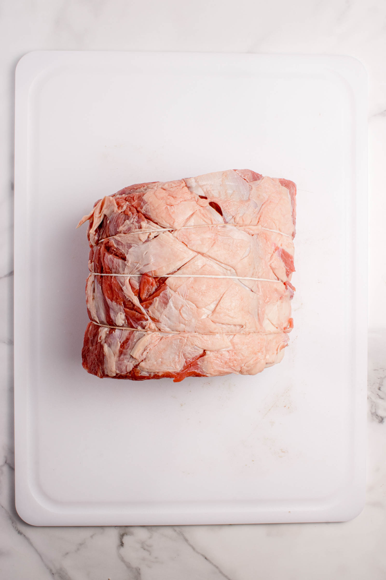 A raw prime rib roast tied with kitchen twine sits on a white cutting board against a marble background.