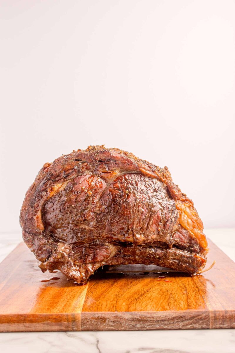 A cooked prime rib roast rests on a wooden cutting board against a plain background.