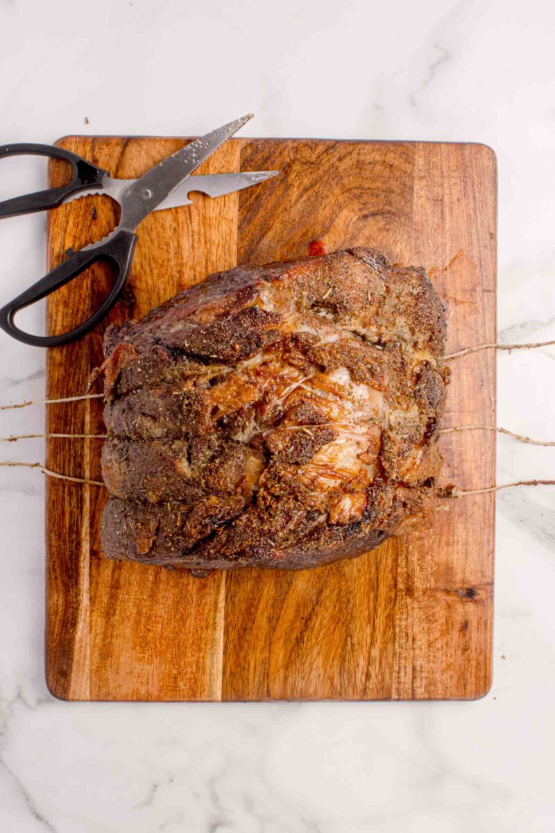 Cooked, seasoned roast beef tied with butcher's twine on a wooden cutting board, with a pair of kitchen shears beside it.
