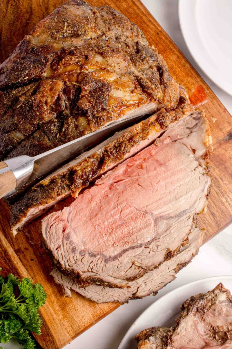 A large piece of seasoned roast beef is being sliced on a wooden cutting board with a knife, showing a juicy pink center.