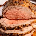 A sliced, seasoned roast beef showing a pink, medium-rare interior on a wooden cutting board with a plate of food and parsley in the background.