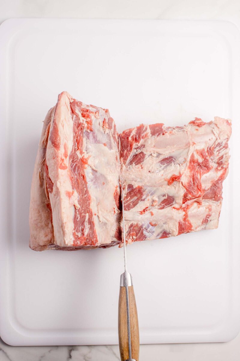 A large piece of raw beef is being cut with a knife on a white cutting board.