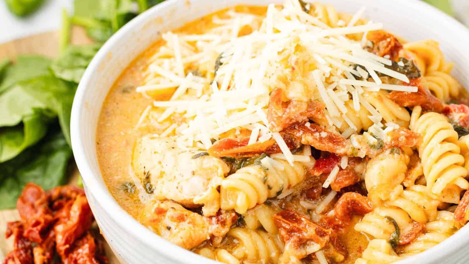 A bowl of creamy pasta with chicken, sun-dried tomatoes, spinach, and shredded cheese on top.
