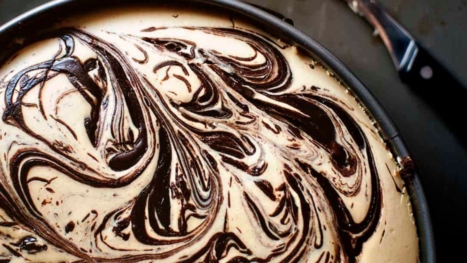 Close-up of a cheesecake with a marbled chocolate swirl pattern on top, in a round baking pan with a knife visible in the background.