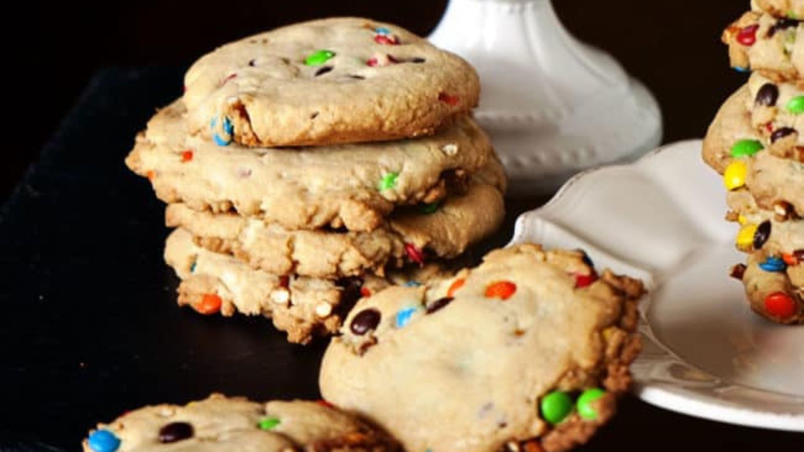 A stack of large, thick cookies with colorful candy-coated chocolates sits next to a white plate holding more cookies.