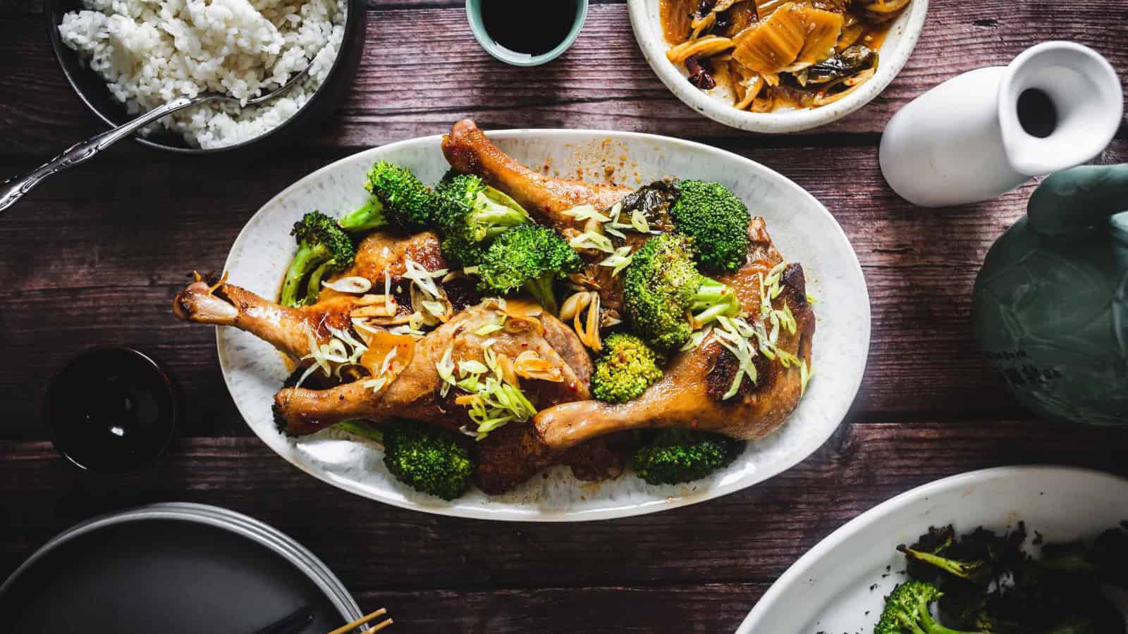 Oval plate of roasted chicken legs with broccoli and scallions, surrounded by rice and side dishes on a wooden table.