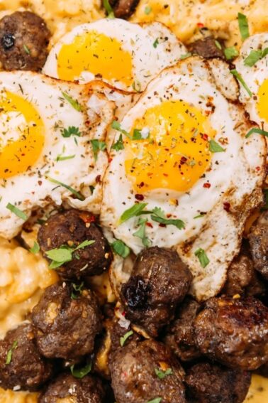 A skillet filled with creamy macaroni and cheese, topped with three sunny-side-up eggs and several browned meatballs, garnished with herbs.