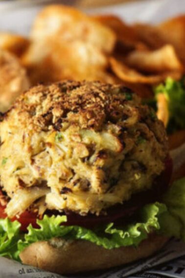 A crab cake sandwich with lettuce and tomato on a bun, served with potato chips and a jar of sauce.