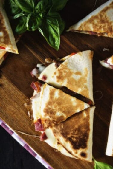 Sliced quesadillas arranged on a wooden board with two bowls of salsa and fresh basil leaves as garnish.