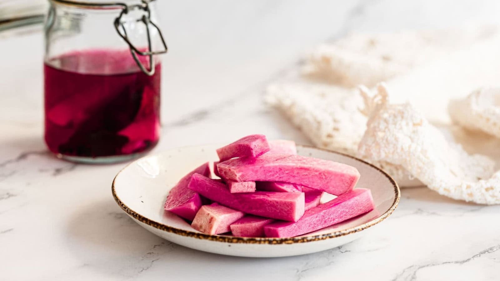 A plate of pink pickled vegetable sticks next to a jar of pink liquid on a marble surface.
