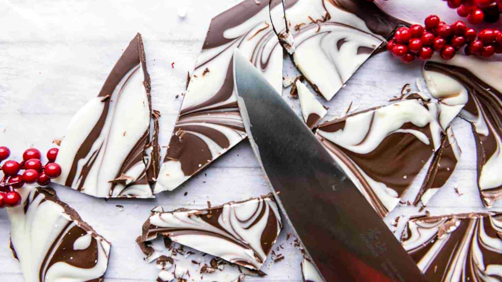 A knife cutting swirled black and white chocolate bark into pieces, with red berries as decoration on the side.