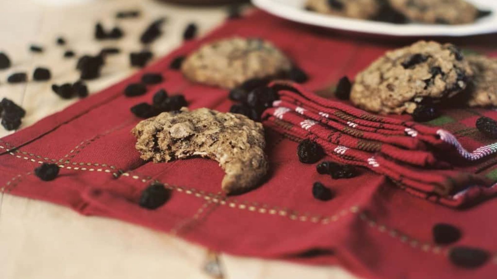 Oatmeal raisin cookies, one with a bite taken out, sit on a red cloth with scattered raisins.
