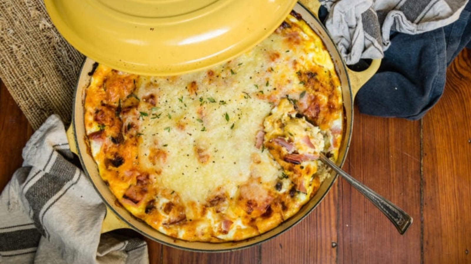 A baked casserole with a golden, cheesy top sits in a round yellow dish, partially covered by a lid, with a serving spoon and kitchen towels nearby.