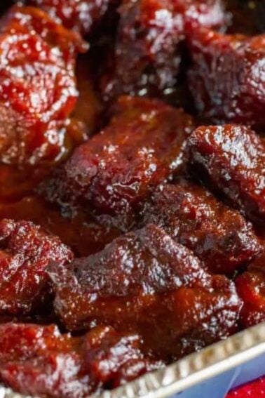 A close-up of saucy, glazed BBQ burnt ends in a foil tray, showing dark, caramelized pieces of meat coated in barbecue sauce.