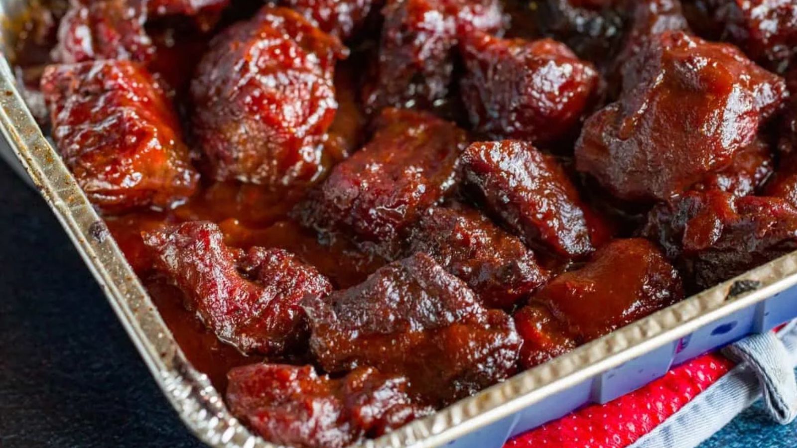A close-up of saucy, glazed BBQ burnt ends in a foil tray, showing dark, caramelized pieces of meat coated in barbecue sauce.