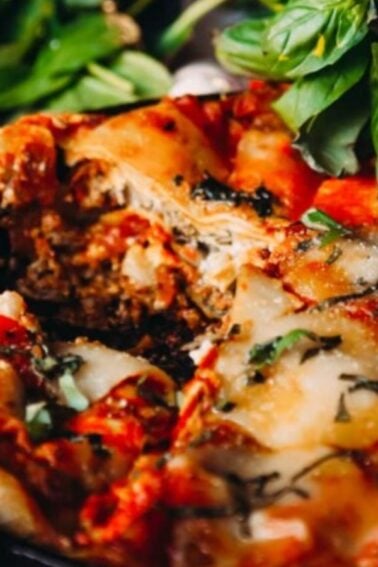 Baked lasagna in a pan topped with melted cheese and fresh basil, with a cluster of tomatoes and leafy greens beside it.