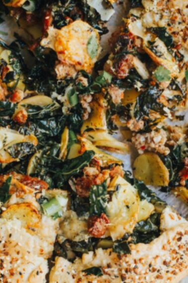 A sliced savory galette topped with leafy greens, cheese, and vegetables on parchment paper, with small yellow potatoes on the side.