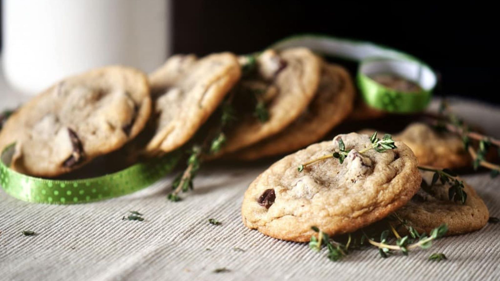 Chocolate chip cookies garnished with fresh herbs are arranged on a cloth, with a green ribbon and a glass of milk in the background.