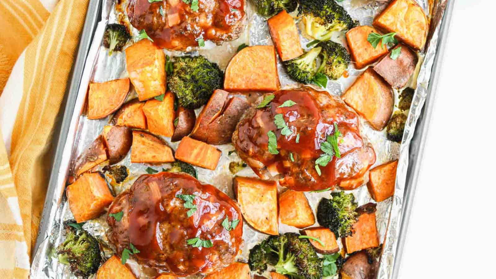 A baking sheet lined with foil holds three glazed meatloaf portions, surrounded by roasted sweet potato cubes and broccoli florets. A yellow and white towel is partially visible to the left.