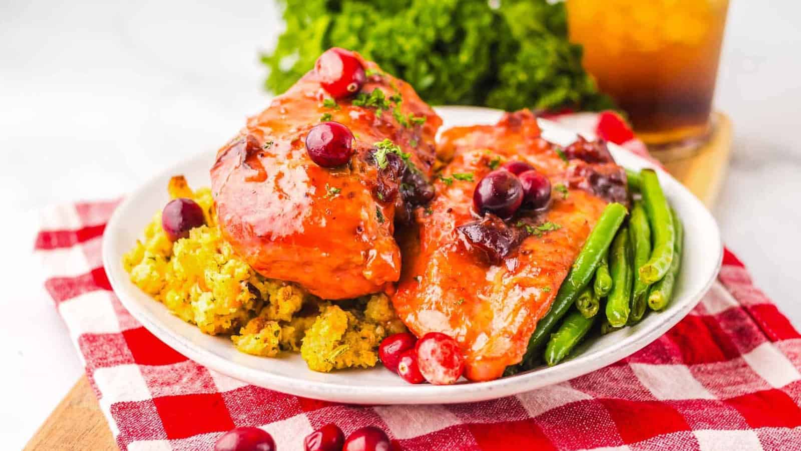 Plate of glazed chicken with cranberries, green beans, and stuffing on a red and white checkered cloth.