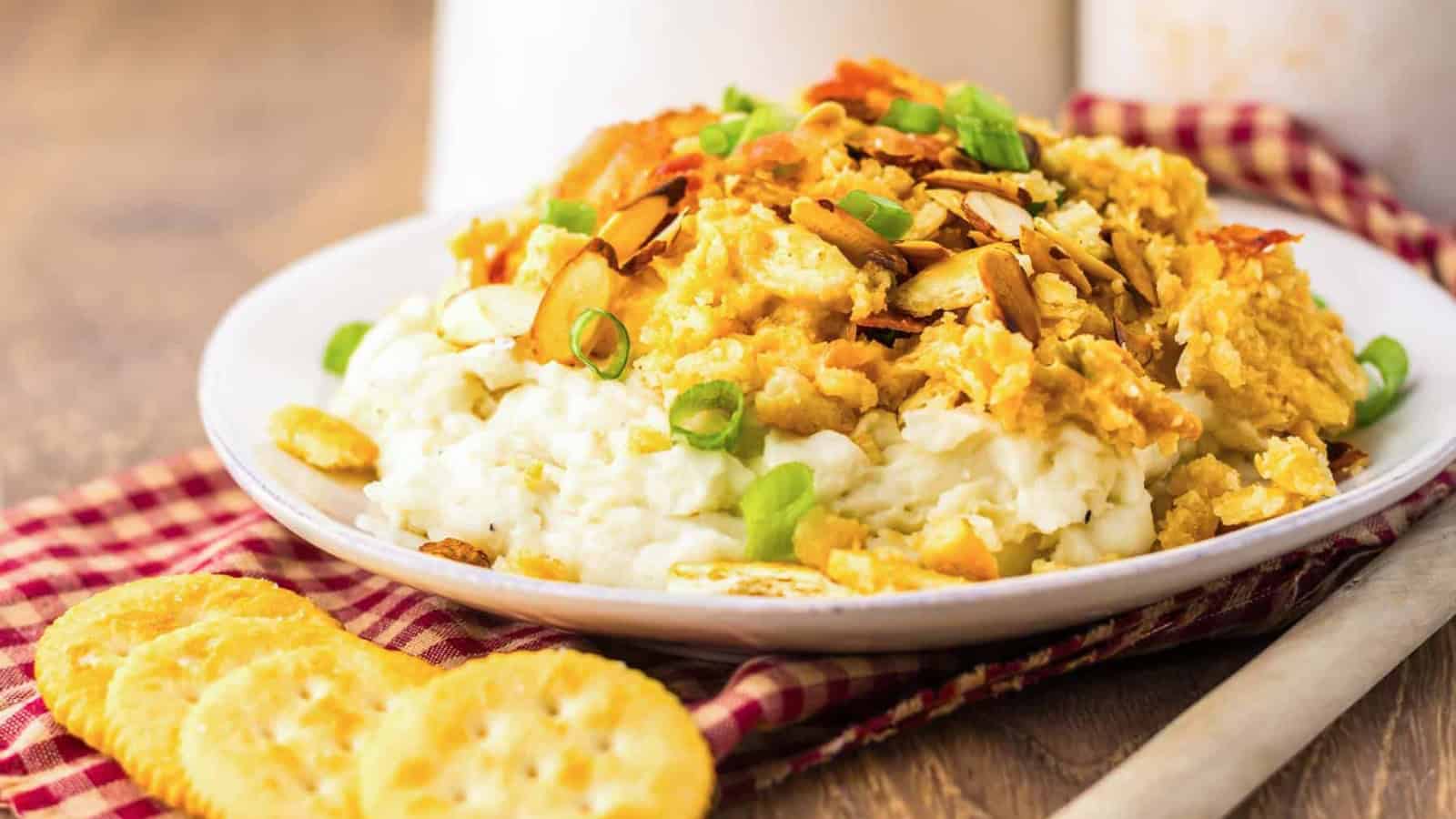 A plate of creamy mashed potatoes topped with a casserole mixture, sliced almonds, green onions, and served with round crackers on the side.