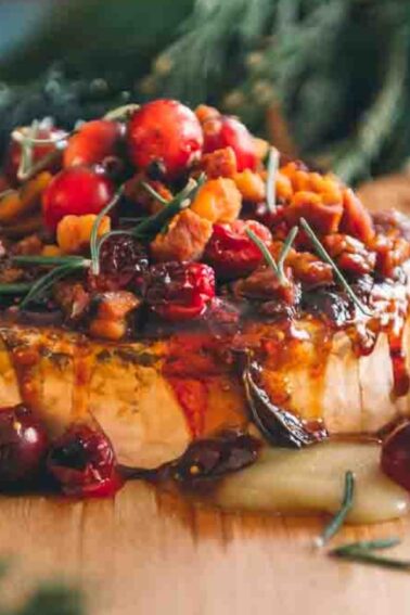 A round baked brie cheese topped with cranberries, nuts, and herbs, served on a wooden board with crackers.