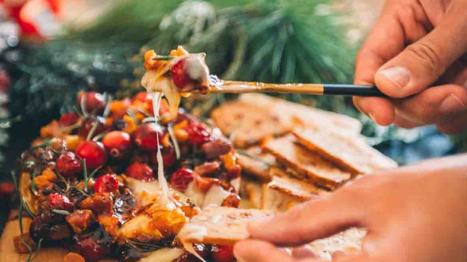 Close-up of hands serving melted cheese topped with cranberries and nuts using a utensil, surrounded by sliced bread on a wooden board.