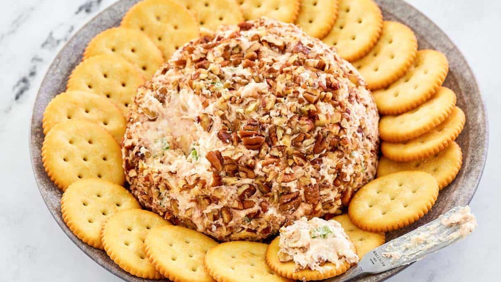 A round cheese ball coated with chopped pecans is served on a plate surrounded by round crackers, with a small spreader on the side.