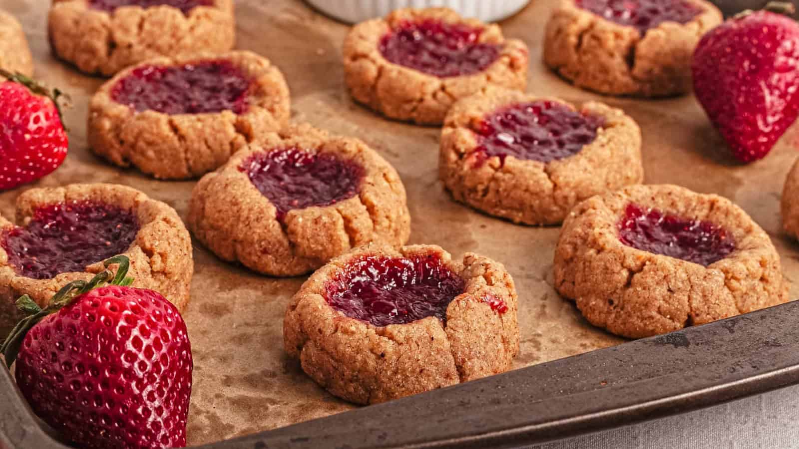 A baking tray with thumbprint cookies filled with red jam, lined with parchment paper, and fresh strawberries placed nearby.