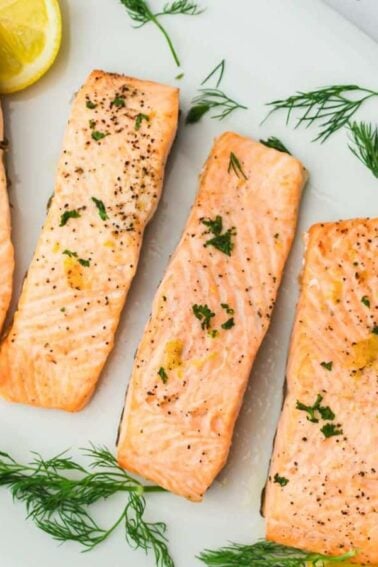 Four cooked salmon fillets garnished with fresh dill and lemon slices are arranged on a white oval plate.