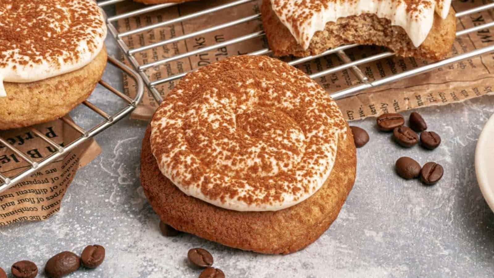 A coffee-flavored cookie topped with cream and cocoa powder sits on a surface with coffee beans nearby; more cookies are on a cooling rack, one with a bite taken out.