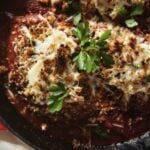 A skillet contains breaded chicken topped with melted cheese and parsley, served over a tomato sauce.