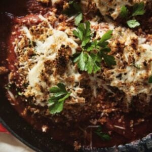 A skillet contains breaded chicken topped with melted cheese and parsley, served over a tomato sauce.
