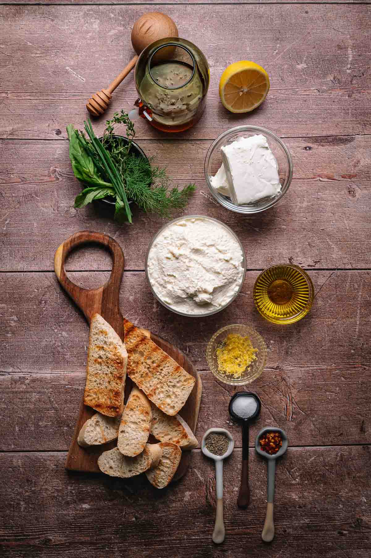 Various ingredients and utensils are arranged on a wooden surface, including bread slices, cheese, herbs, lemon, honey, oil, and small bowls of spices.