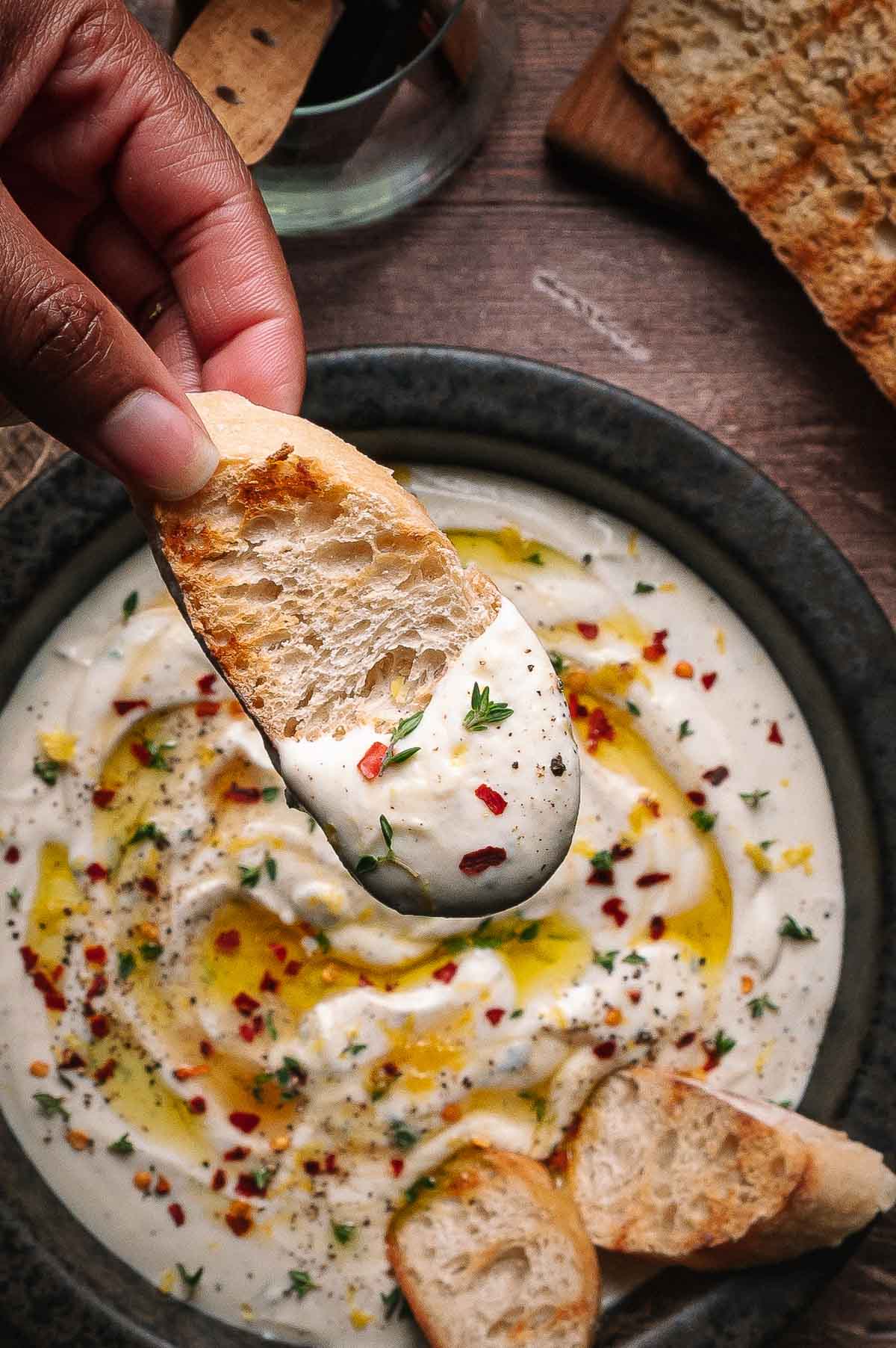 A hand dips a toasted bread slice into a bowl of creamy whipped ricotta, garnished with olive oil, red pepper flakes, herbs, and served with more bread slices.