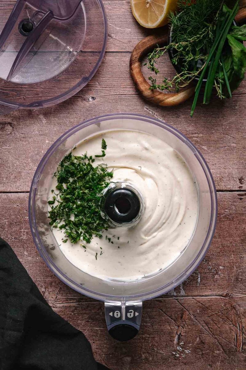 A food processor containing creamy white whipped ricotta with chopped fresh herbs on top, placed on a wooden surface next to fresh herbs and a lemon half.