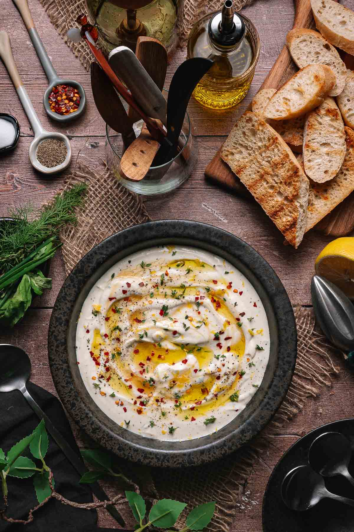 A bowl of creamy whipped ricotta dip garnished with herbs, olive oil, and red pepper flakes, surrounded by sliced bread, fresh herbs, and condiments on a rustic wooden table.