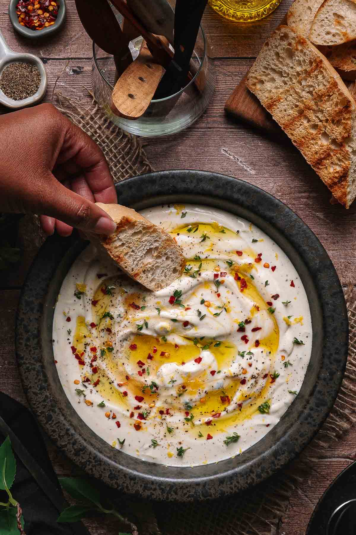 A hand dips a piece of bread into a bowl of creamy whipped ricotta dip topped with olive oil, herbs, and chili flakes. Toasted bread slices are on the side.