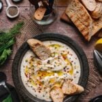 A bowl of creamy whipped ricotta garnished with olive oil, herbs, and red pepper flakes, served with toasted bread slices on a rustic wooden table with various kitchen tools nearby.