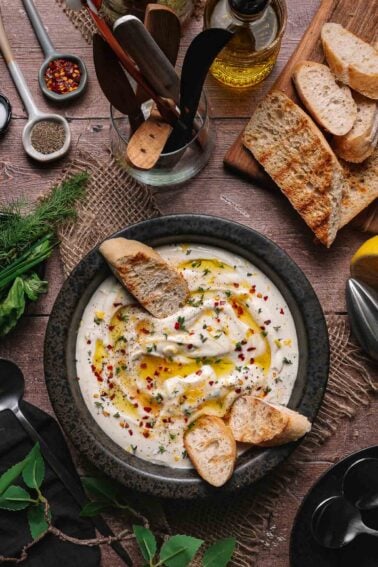A bowl of creamy whipped ricotta garnished with olive oil, herbs, and red pepper flakes, served with toasted bread slices on a rustic wooden table with various kitchen tools nearby.