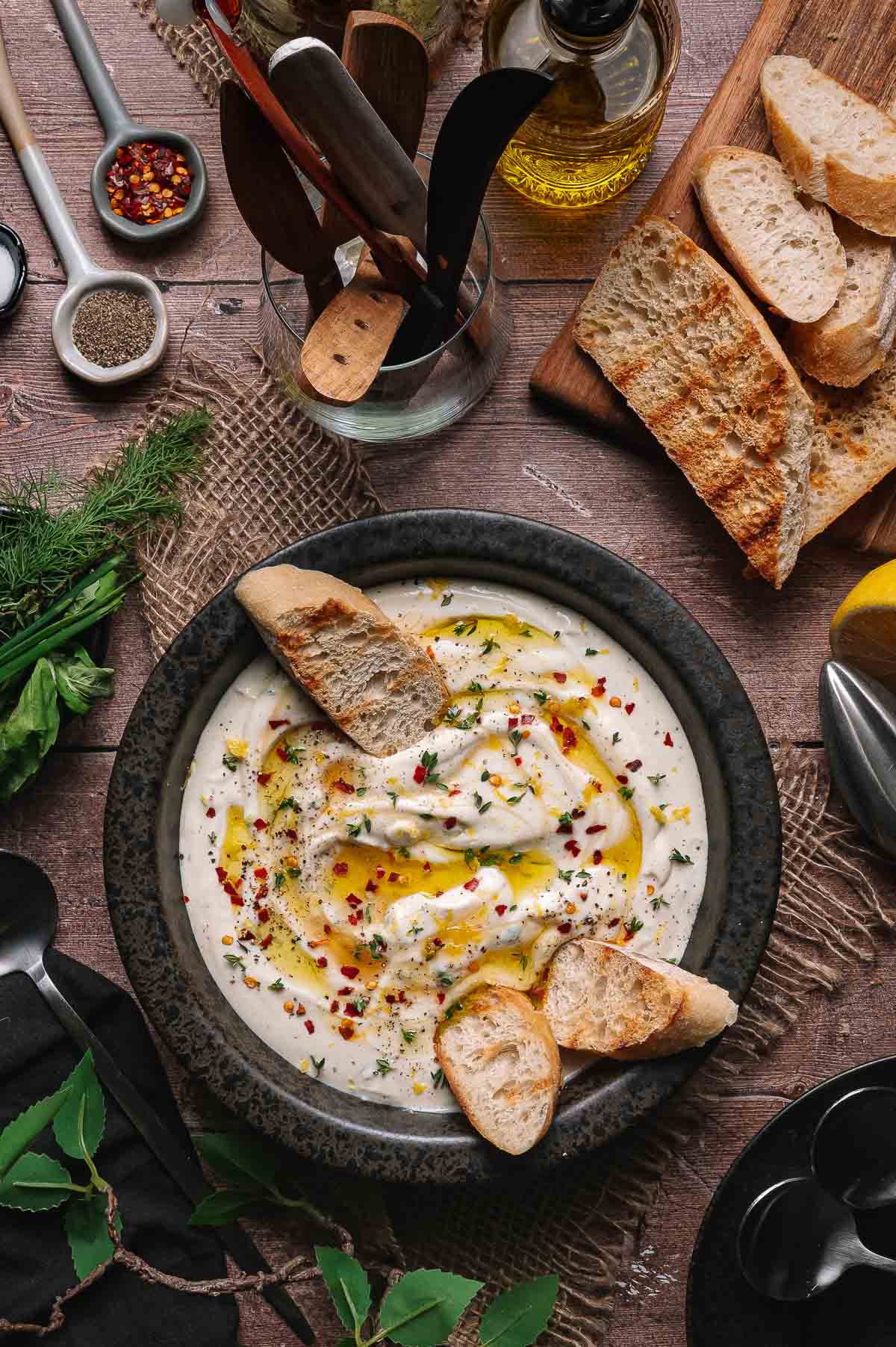 A bowl of creamy whipped ricotta garnished with olive oil, herbs, and red pepper flakes, served with toasted bread slices on a rustic wooden table with various kitchen tools nearby.