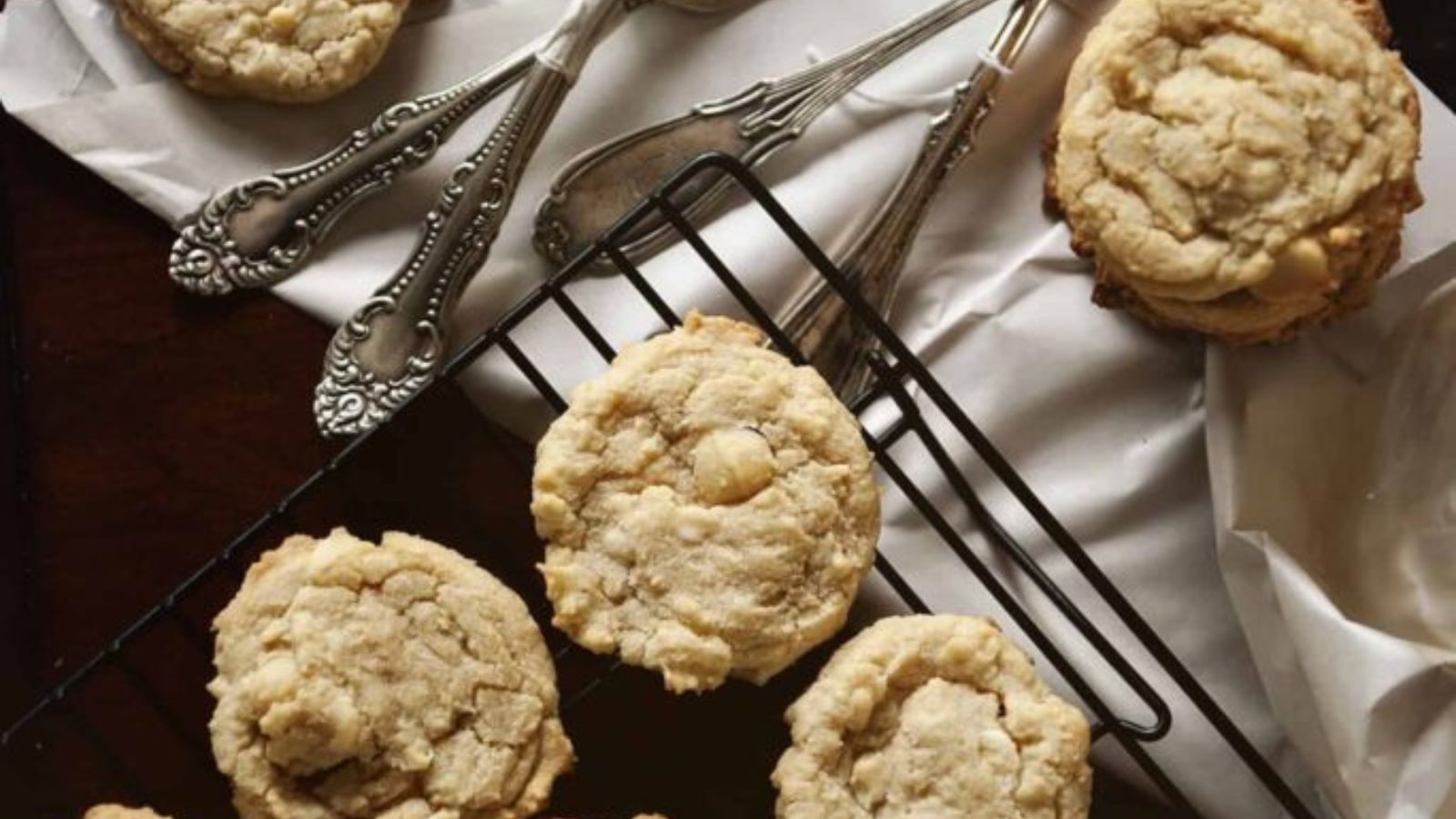 A cooling rack and parchment paper with several cookies and vintage silverware arranged on a dark wooden surface.