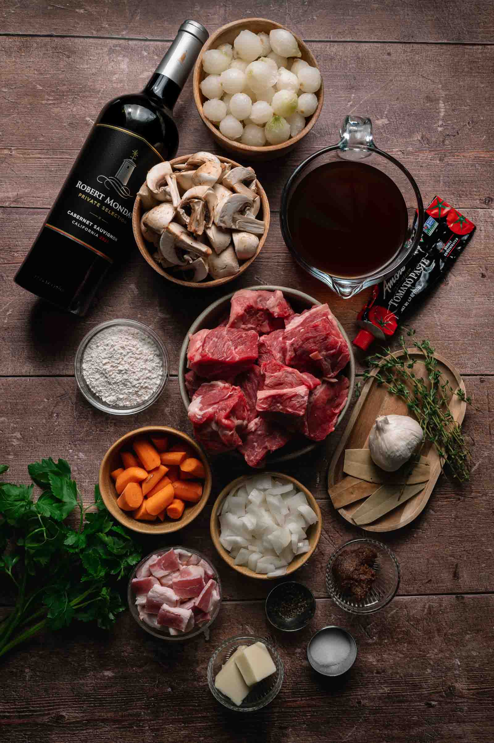 An overhead view of ingredients for beef bourguignon, including wine, beef, onions, mushrooms, carrots, bacon, herbs, garlic, butter, flour, tomato paste, and broth.