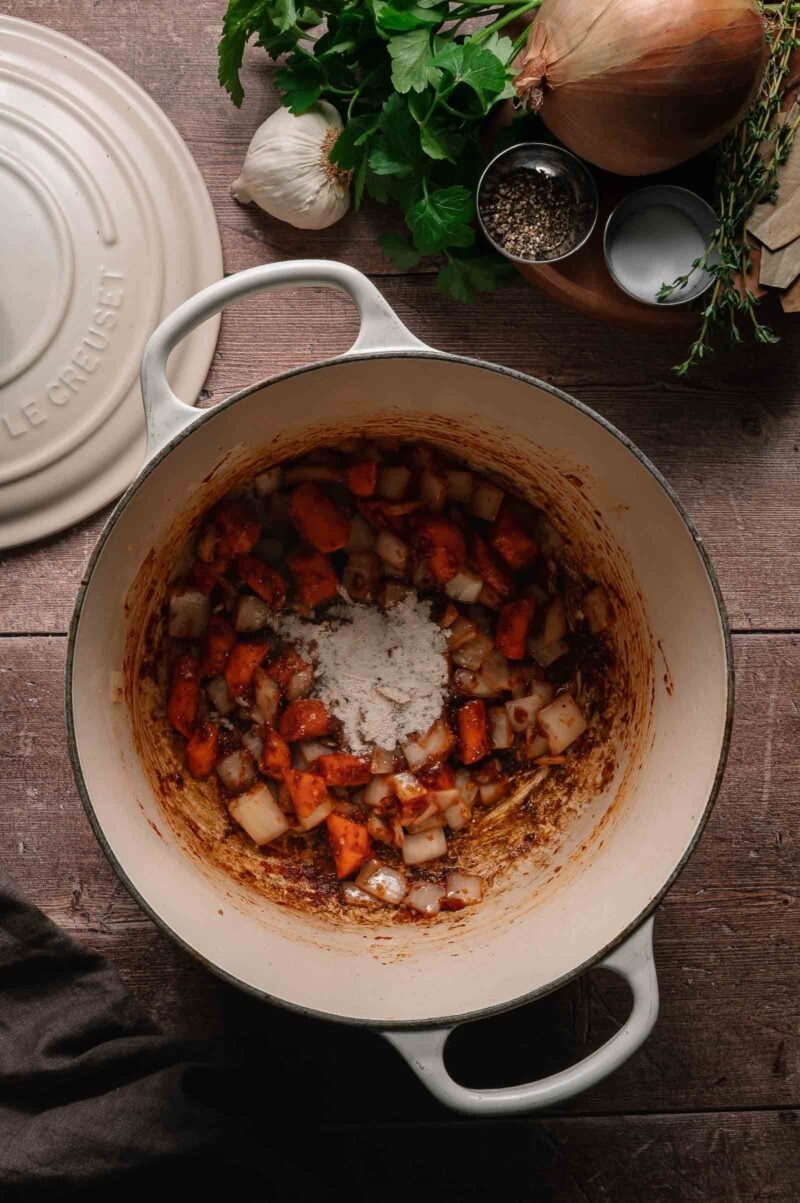 Chopped carrots and onions cooking in a white Dutch oven, sprinkled with flour, surrounded by herbs, spices, and a garlic bulb on a wooden surface.