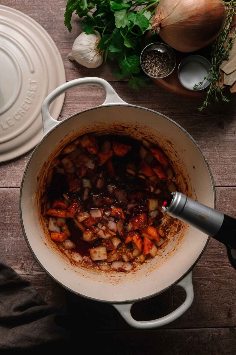 Chopped onions and carrots are sautéing in a white Dutch oven, while red wine is being poured in; herbs, garlic, onion, and pepper are arranged nearby.
