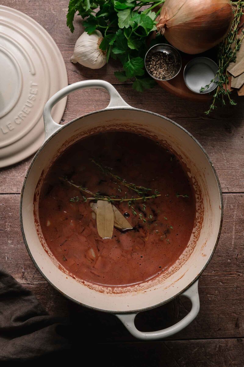A white Dutch oven filled with simmering brown stew, garnished with bay leaves and thyme, sits on a wooden surface beside herbs, onion, garlic, spices, and a pot lid.