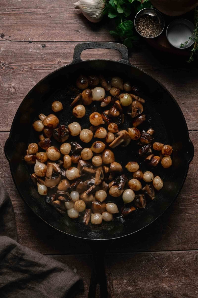 A cast iron skillet filled with sautéed mushrooms and pearl onions, placed on a wooden surface with herbs, garlic, and pepper nearby.